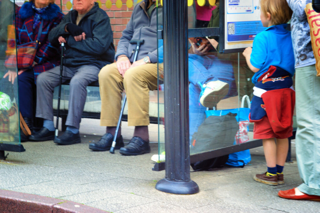 Pensioners waiting at a bus shelter with a young boy reading the departure board with their parent behind them. It is a low angle view looking at the scene at a child's eye level.
